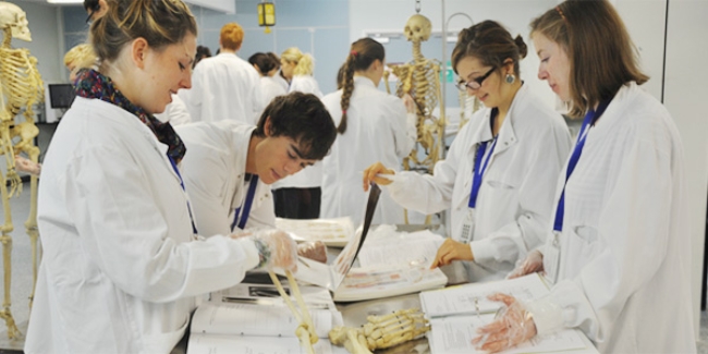 Four anatomy students stand around a table in a laboratory, on the table there are a number of academic textbooks and a model of the bones in a human leg. In the background other students are studying models of human skeletons.
